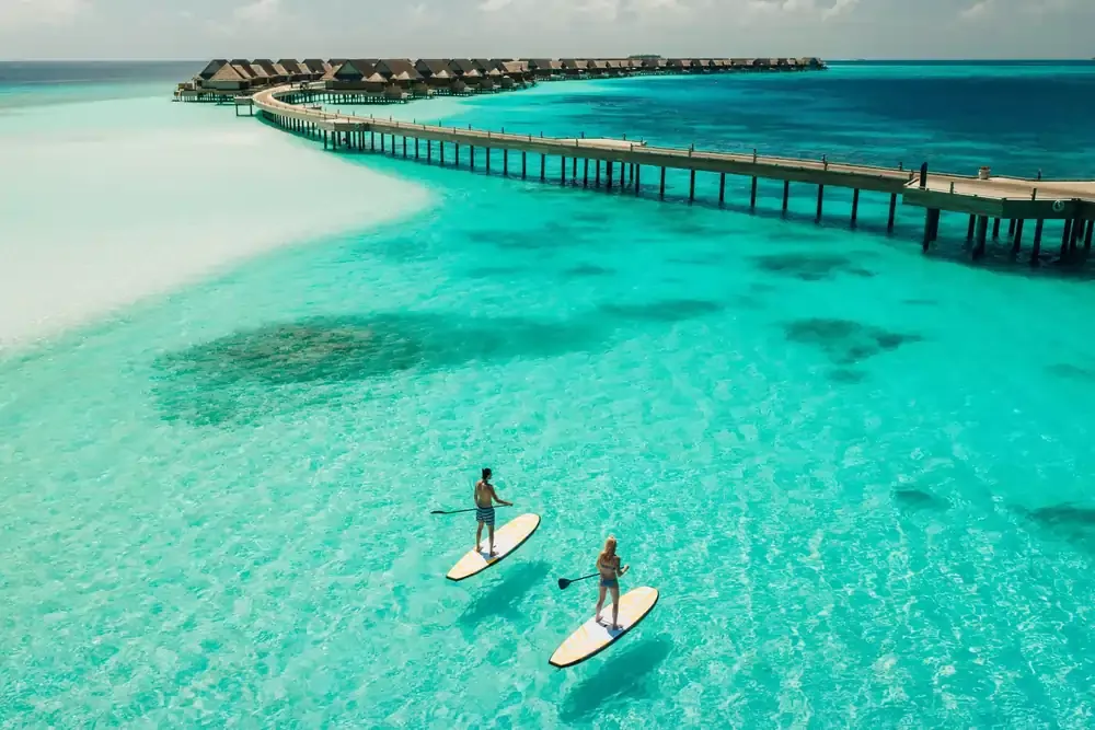 Couple paddleboarding on clear turquoise waters in the Maldives, near a luxury overwater resort with thatched-roof villas connected by a long wooden walkway.