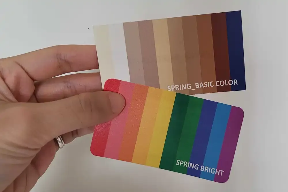 Hand holding two personal color analysis cards labeled "SPRING_BASIC COLOR" with neutral shades and "SPRING BRIGHT" featuring a rainbow of vibrant colors, used during a personal color consultation in Seoul.