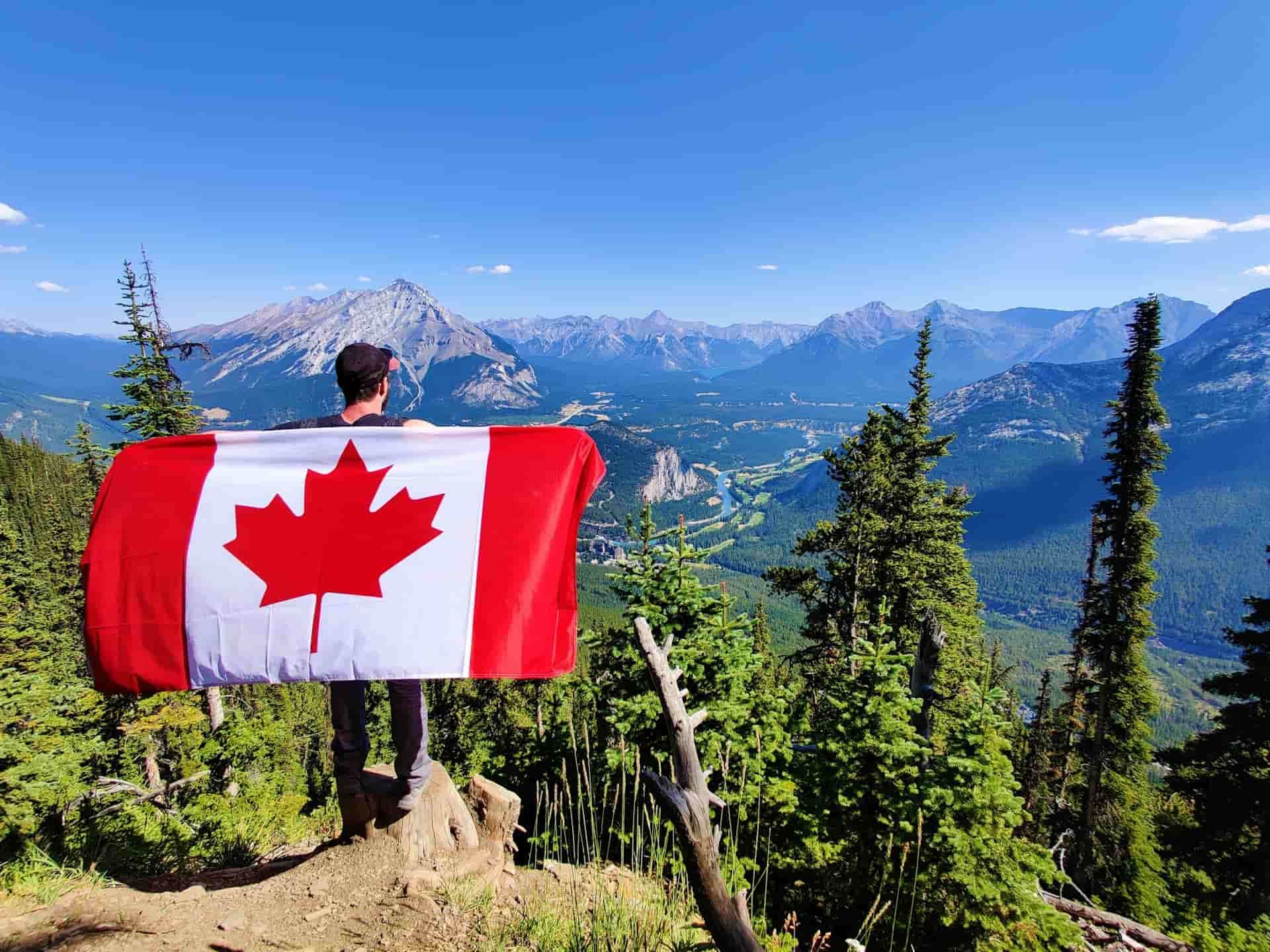 A hiker holding a Canadian flag on a mountain peak overlooking a lush valley in Banff National Park.