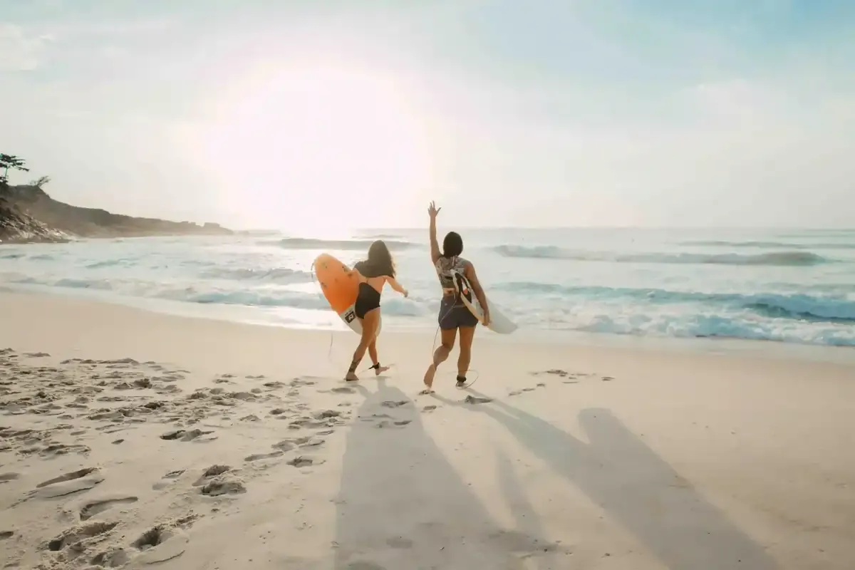Two women running into the ocean with surfboards during a sunny beach day, capturing the carefree spirit of a spring break escape.