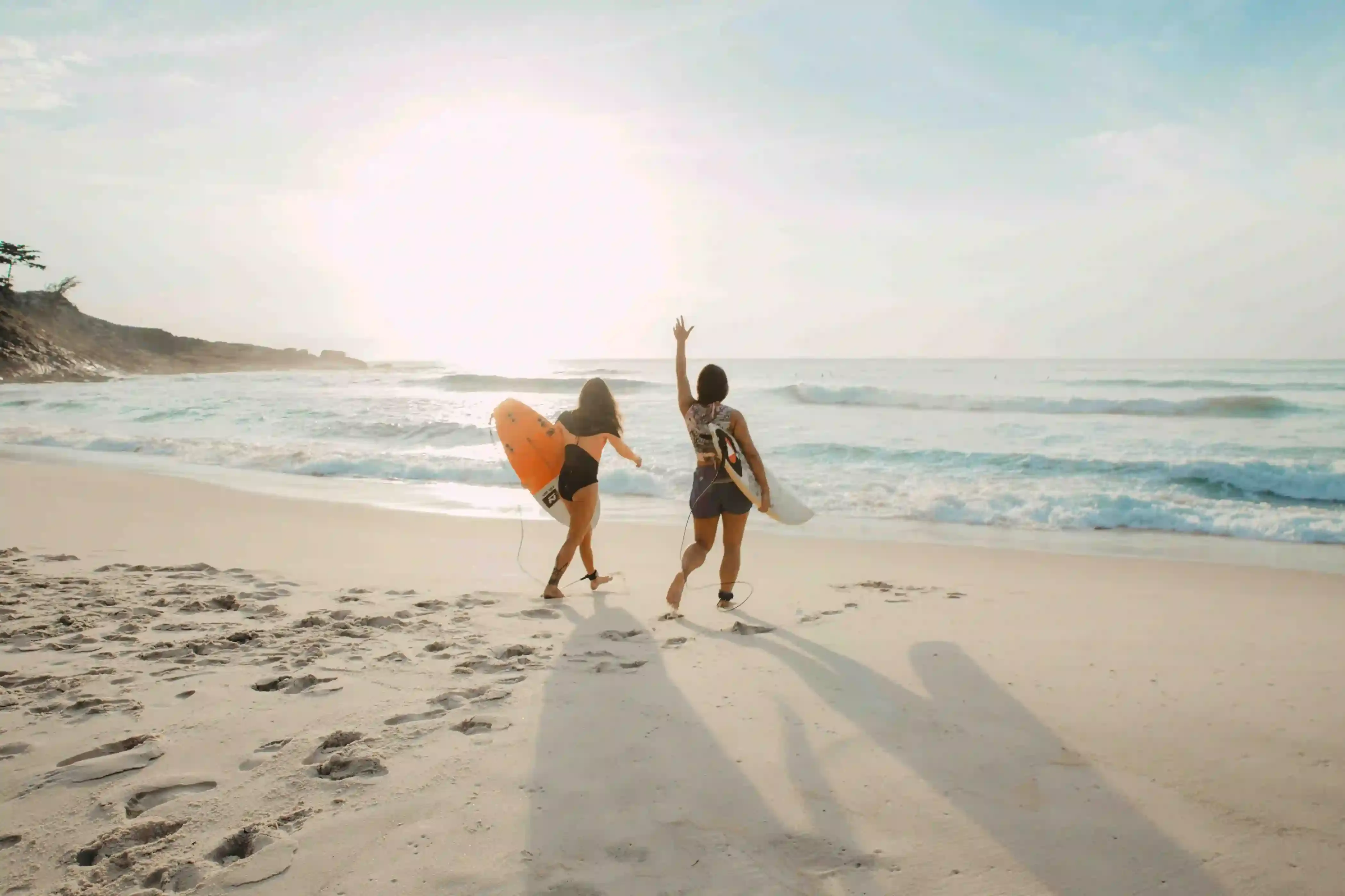 Two women running into the ocean with surfboards during a sunny beach day, capturing the carefree spirit of a spring break escape.