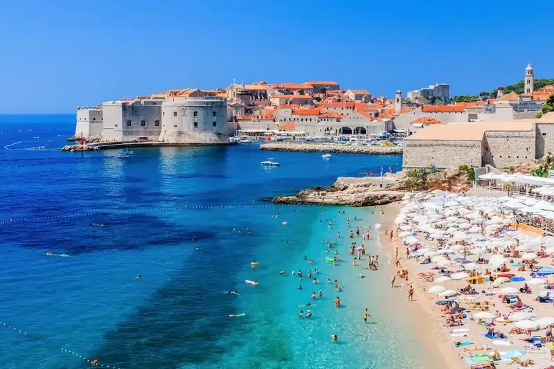 Banje Beach in Dubrovnik, Croatia with sunbathers, turquoise Adriatic waters, and the historic Old Town with medieval walls and terracotta rooftops in the background.