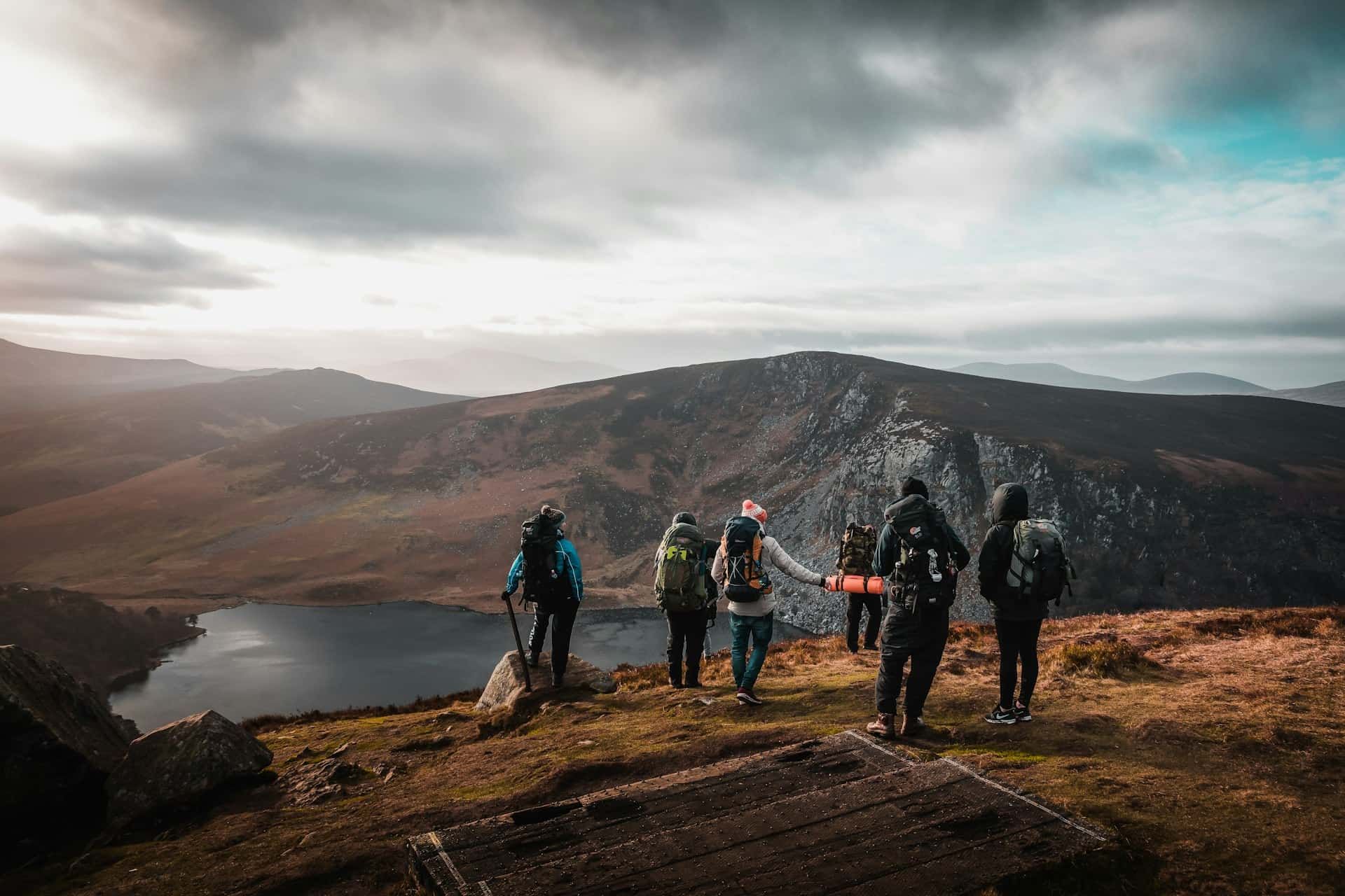 A group of six hikers with large backpacks standing on a rugged mountain ridge overlooking a dark valley lake during a European backpacking trip