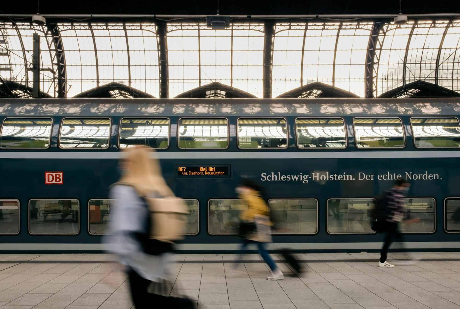  Backpackers with luggage walking briskly past a Deutsche Bahn train on a station platform in Germany.