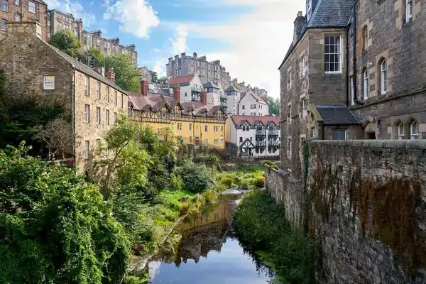 Charming view of Dean Village along the Water of Leith in Edinburgh, Scotland, with stone houses, lush greenery, and a tranquil stream running through the historic neighborhood.