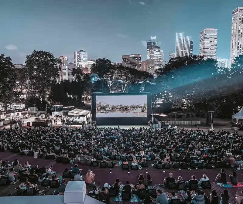 Large crowd enjoying an outdoor movie screening at Film at the Fort in Singapore, with city skyline and trees framing the open-air cinema experience at dusk.