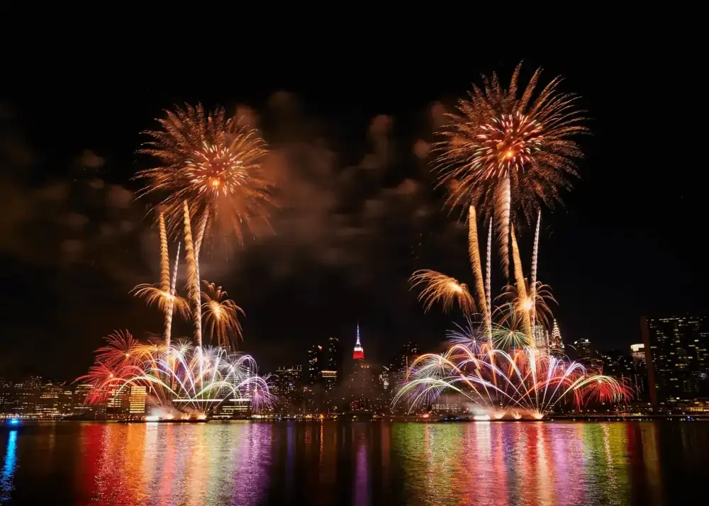 July 4th fireworks lighting up the New York City night sky with rainbow-colored reflections on the water.