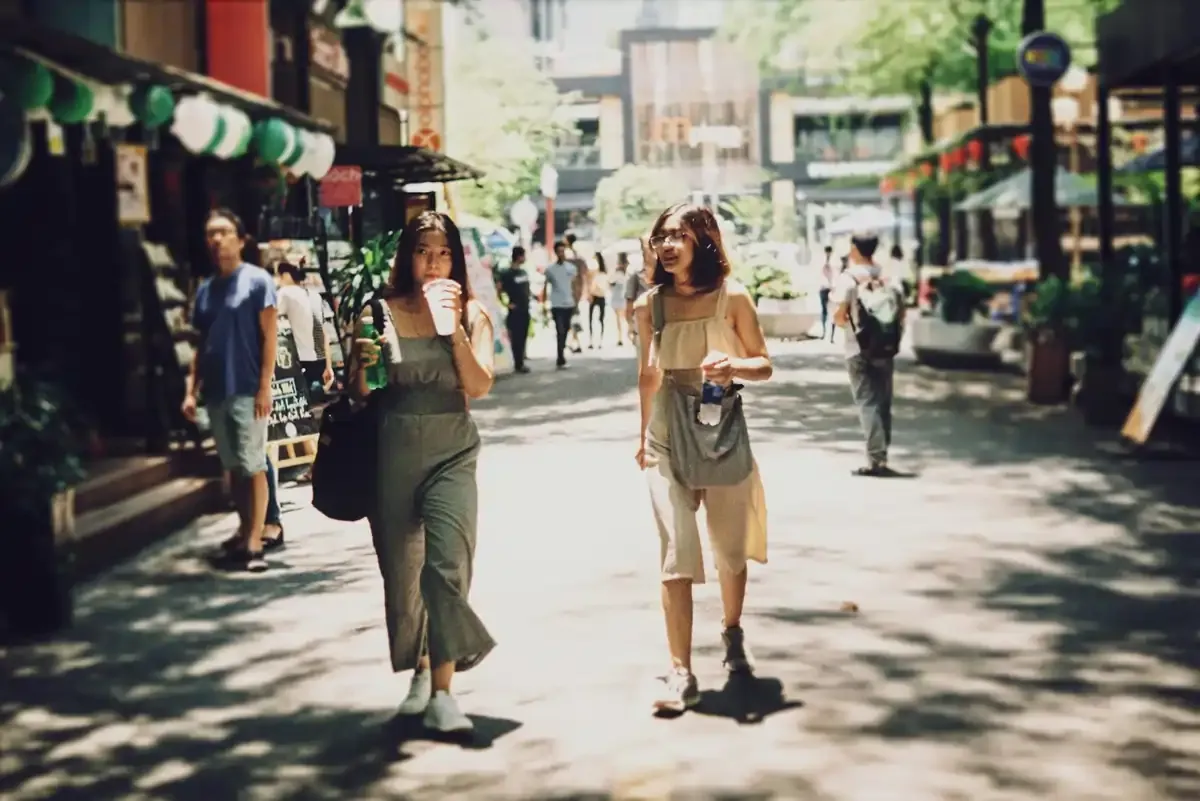 Two women walking along a sunny shopping street in Singapore, sipping drinks and enjoying a casual day out.