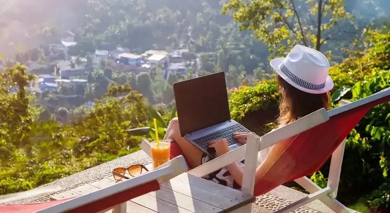 Digital nomad woman working remotely on a laptop from a scenic tropical location, with a village view and a refreshing drink by her side.