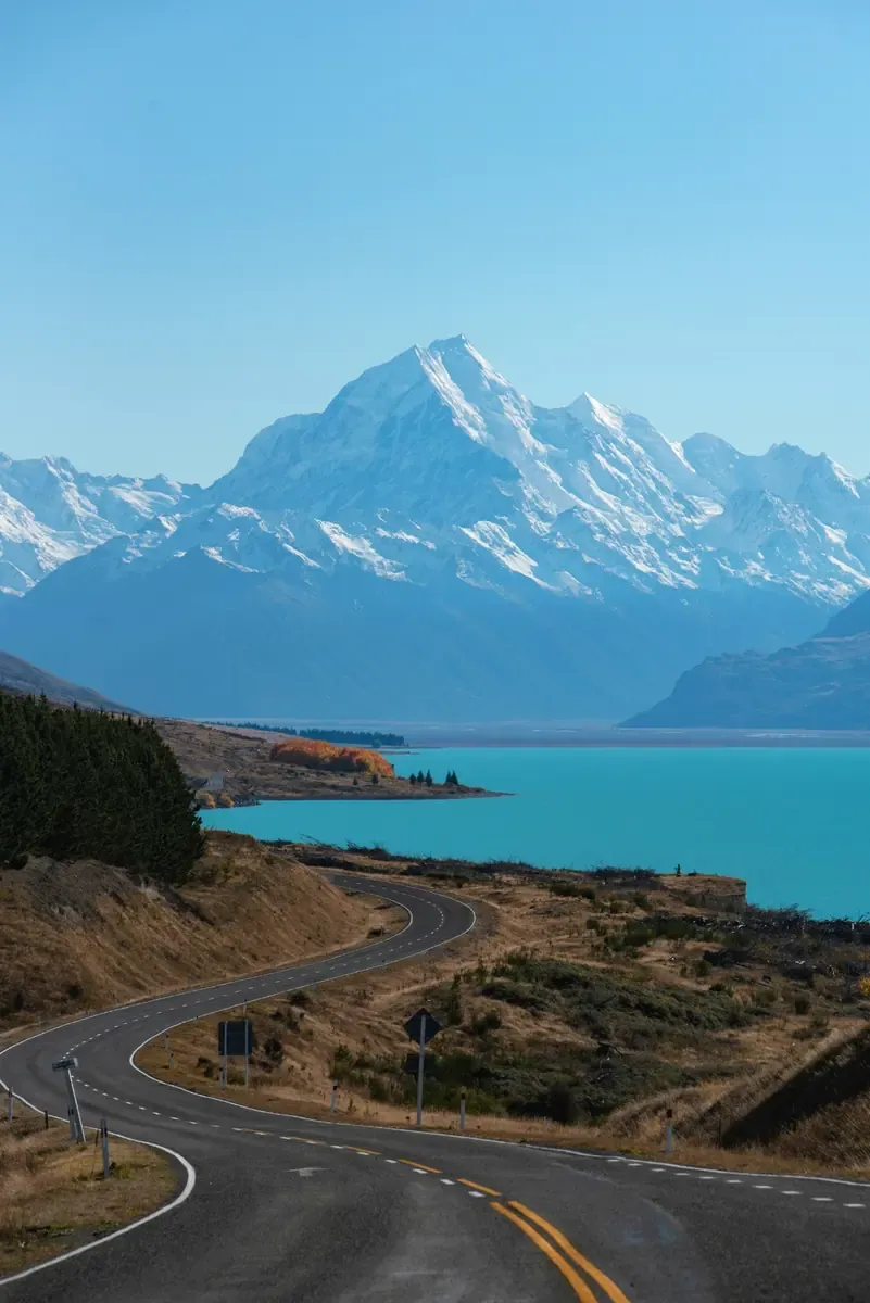 Winding road beside a turquoise lake with snow-covered Aoraki Mount Cook in the distance.