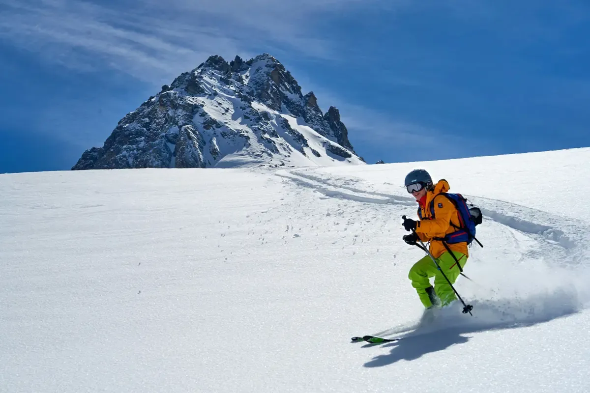 Skier descending a snowy Alpine slope with a rugged mountain peak in the background.