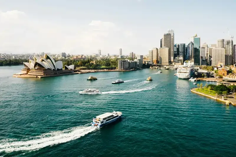 Sydney skyline with the Opera House, boats on blue waters, and lush green waterfront across Sydney Harbor.