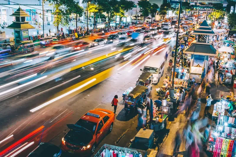 Bangkok street scene at night with light trails from traffic, roadside stalls, and crowds moving through the city.
