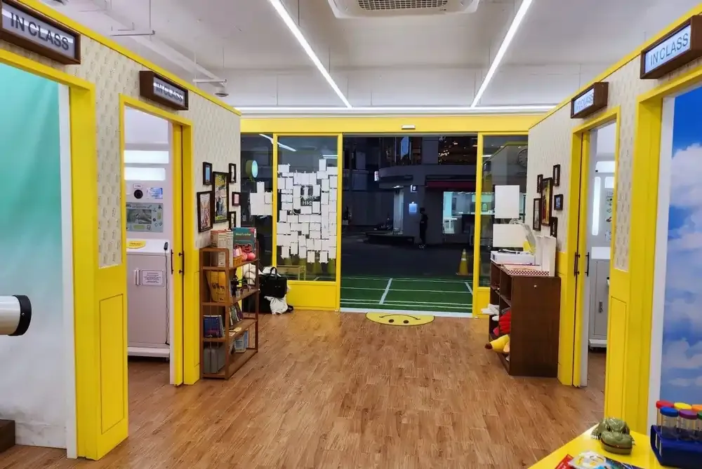 Brightly lit entrance of a self-photo studio in Seoul featuring yellow-framed rooms labeled “IN CLASS,” with wooden flooring and decor evoking a playful classroom setting.