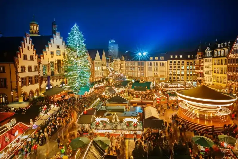 Frankfurt Christmas Market in Germany with festive stalls, carousel, and large decorated Christmas tree at night.