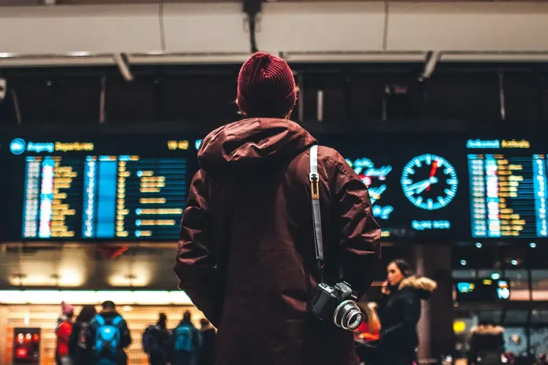 Traveler looking up at a large airport departure board, with a camera strapped over their shoulder.