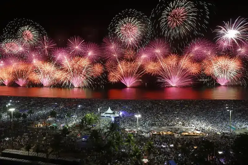 Massive crowd gathered on a beach watching vibrant fireworks light up the night sky over the water during a New Year's Eve celebration in Rio de Janeiro, Brazil.