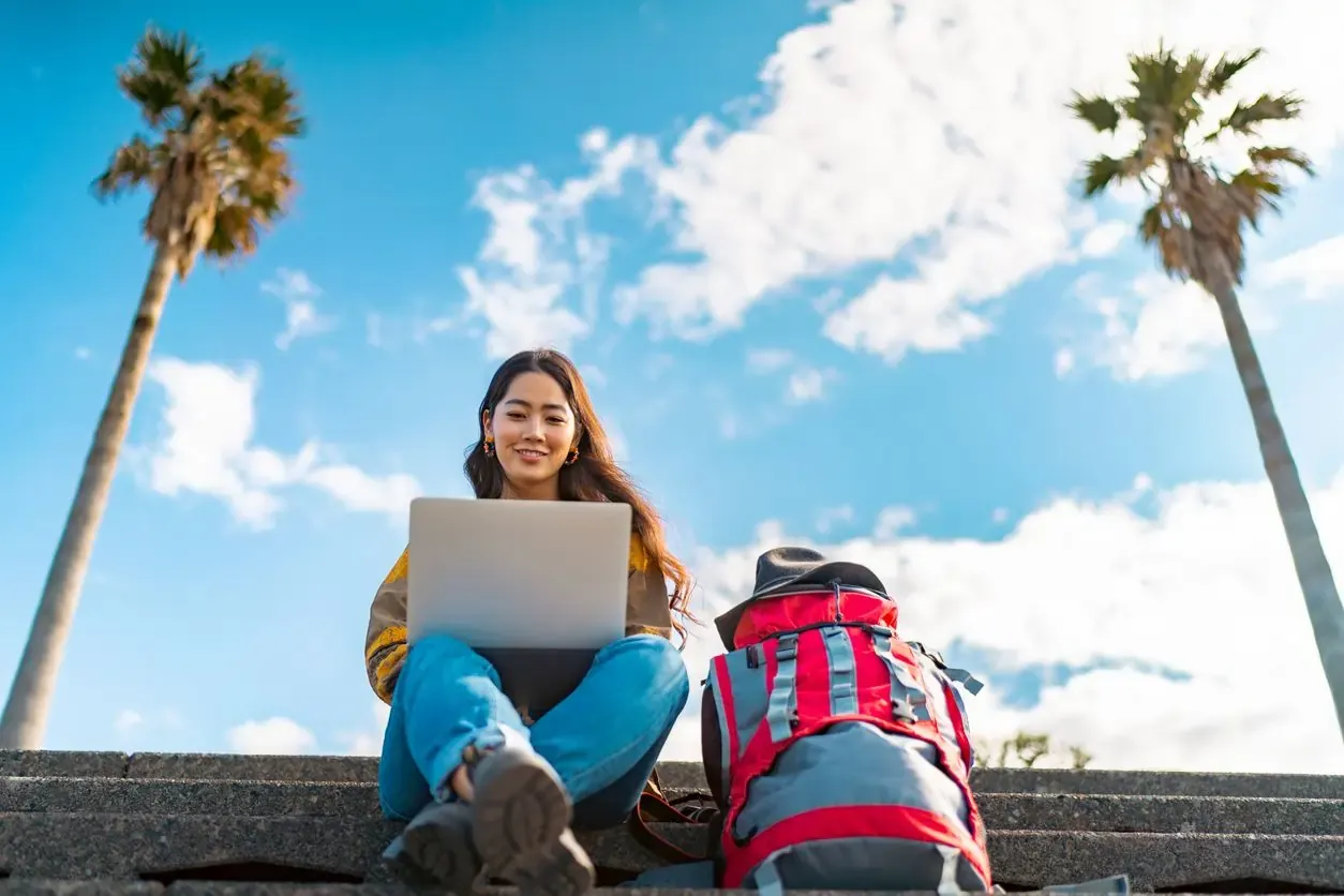 Smiling traveler working on a laptop outdoors under blue skies with palm trees, showing how eSIMs enable internet access on the go.