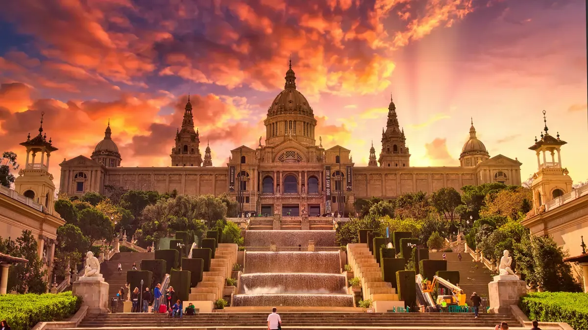Palau Nacional in Barcelona illuminated at sunset, overlooking Montjuïc fountains and terraces with dramatic orange clouds.