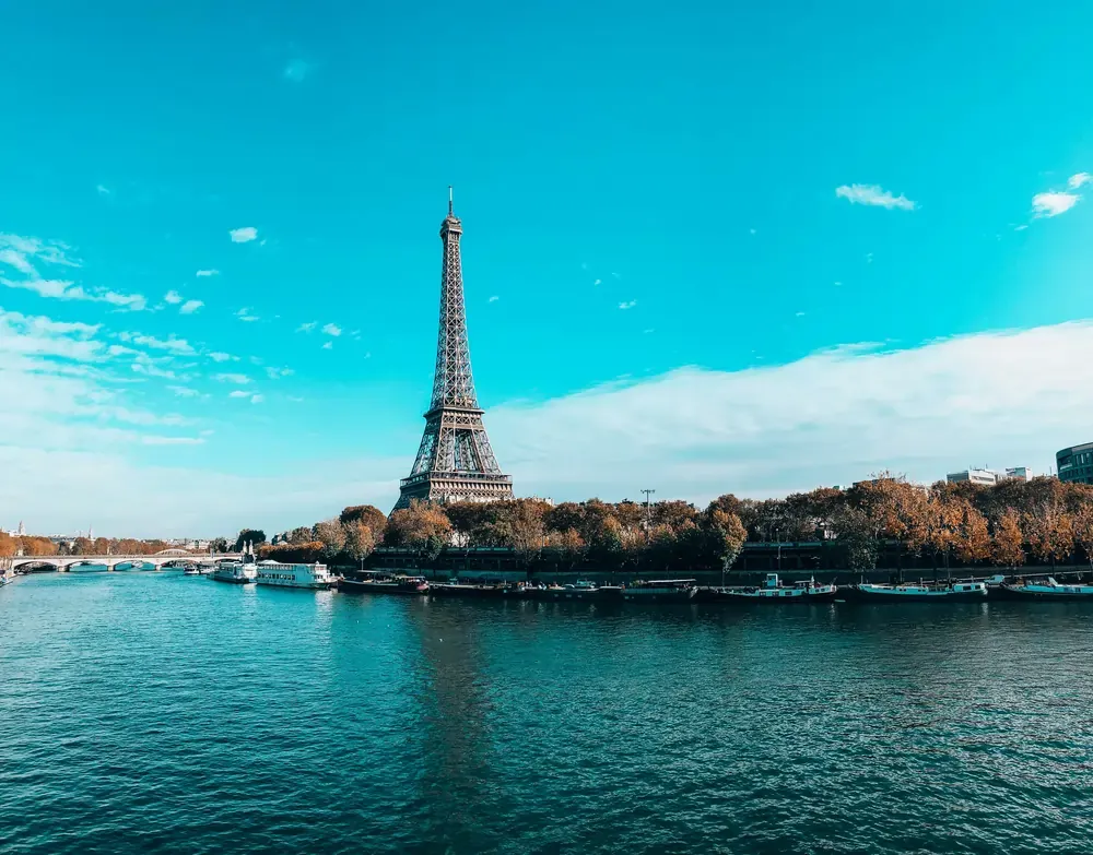 Eiffel Tower overlooking the River Seine in Paris on a bright day, with boats and tree-lined riverbanks.