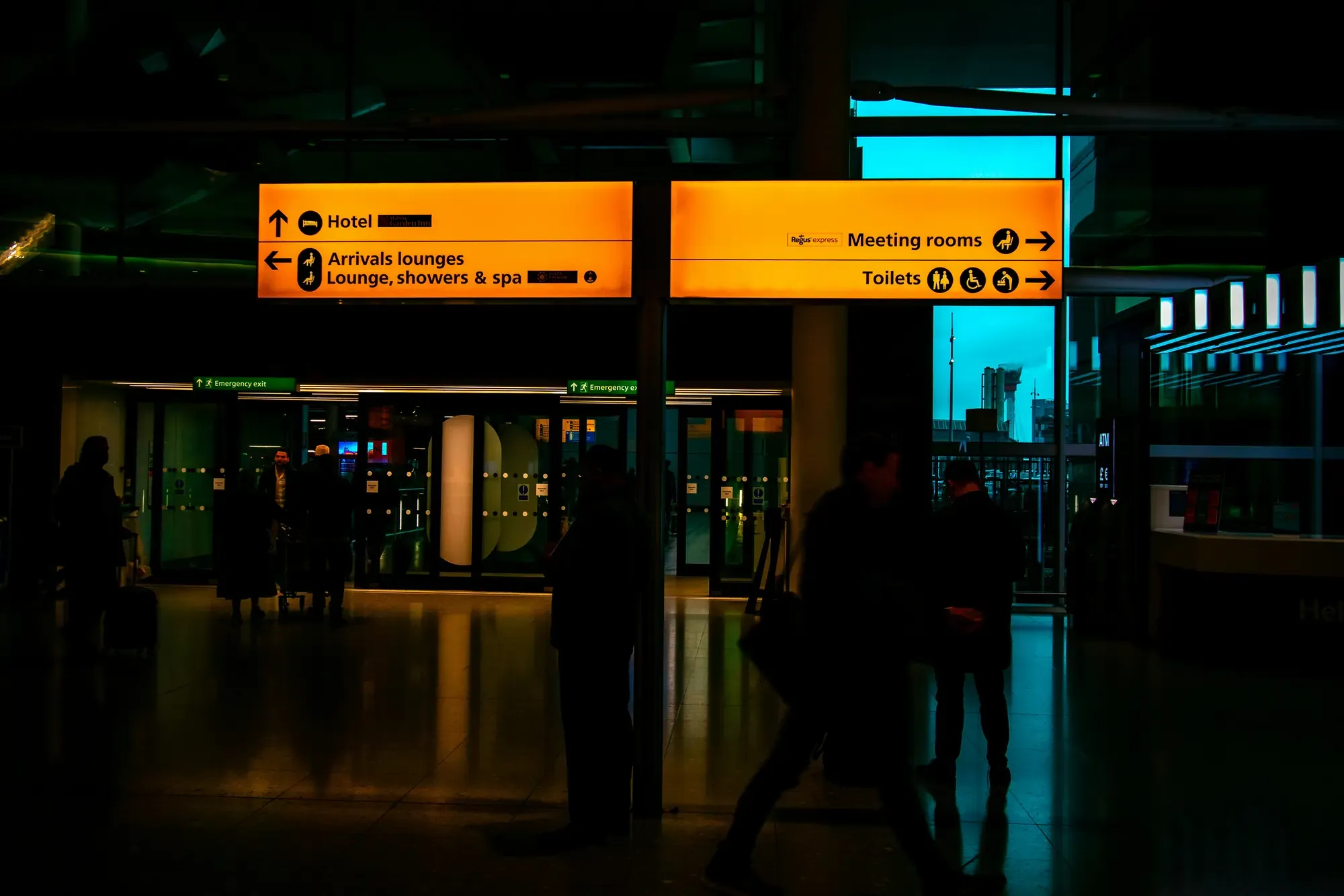 Travelers walking beneath directional signs inside a London airport arrivals area.