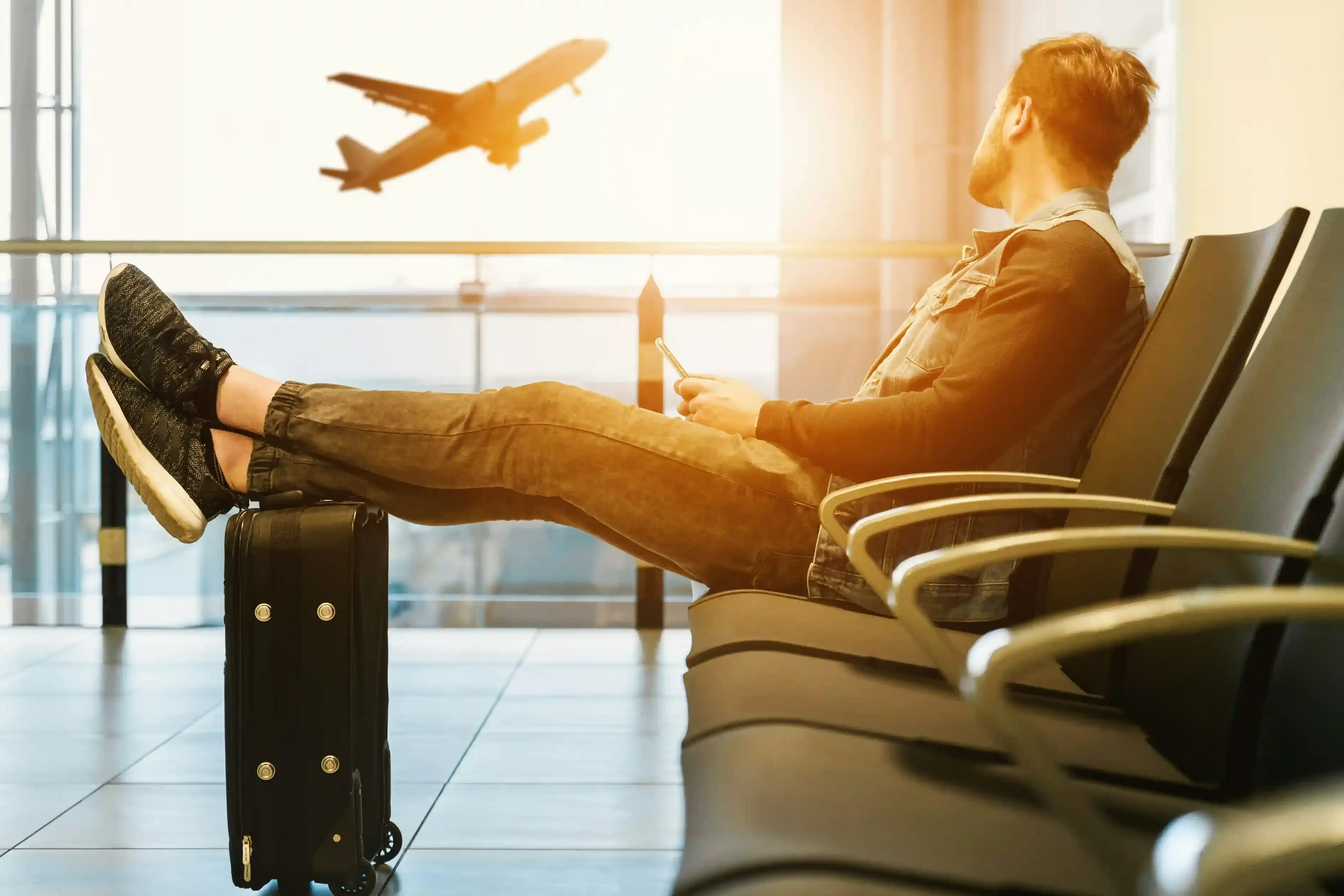 Traveler at airport lounge using smartphone with legs resting on suitcase, watching airplane take off at sunrise – representing seamless mobile connectivity while traveling.