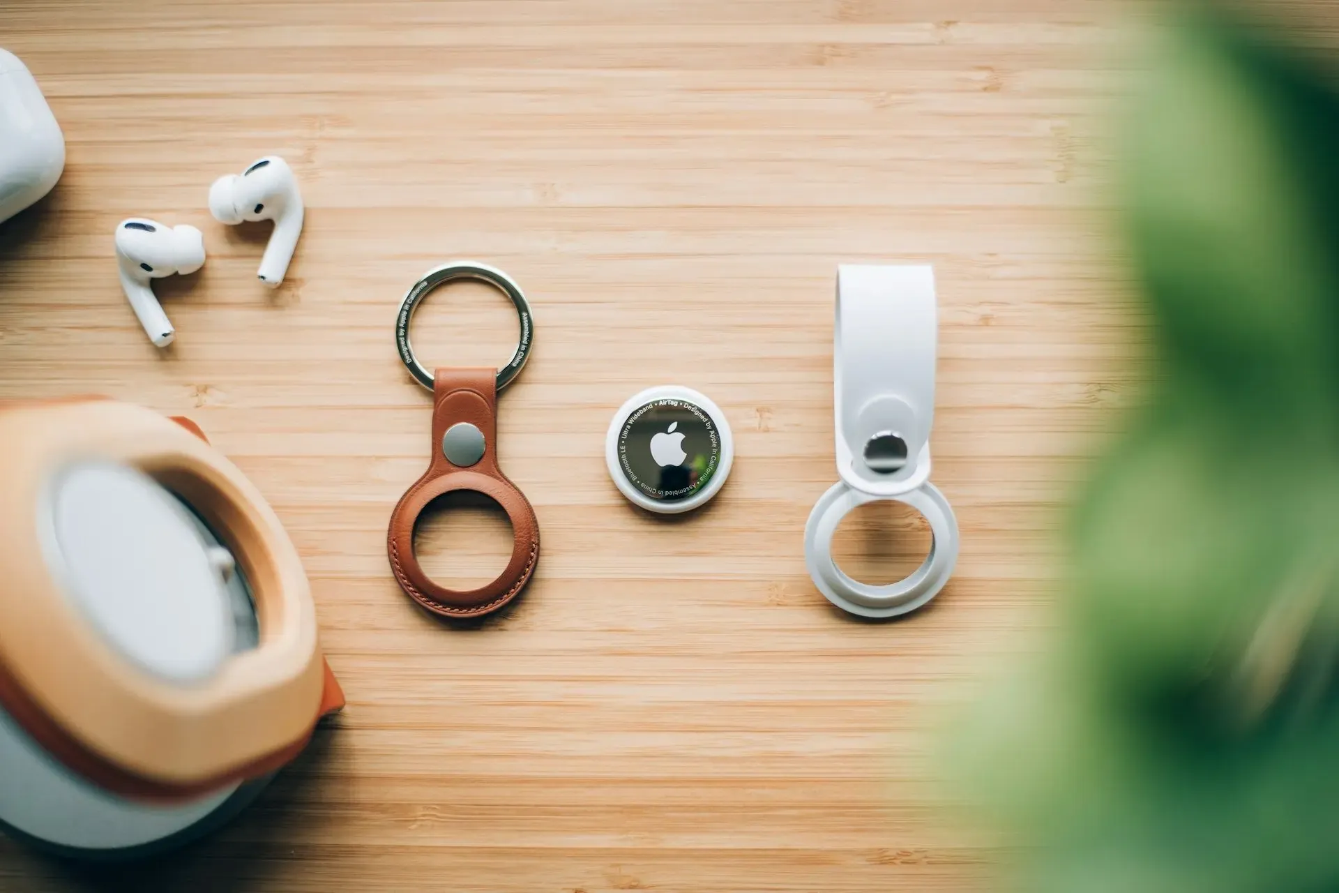 Flat lay of Apple accessories including an AirTag, leather and plastic holders, and AirPods on a wooden surface