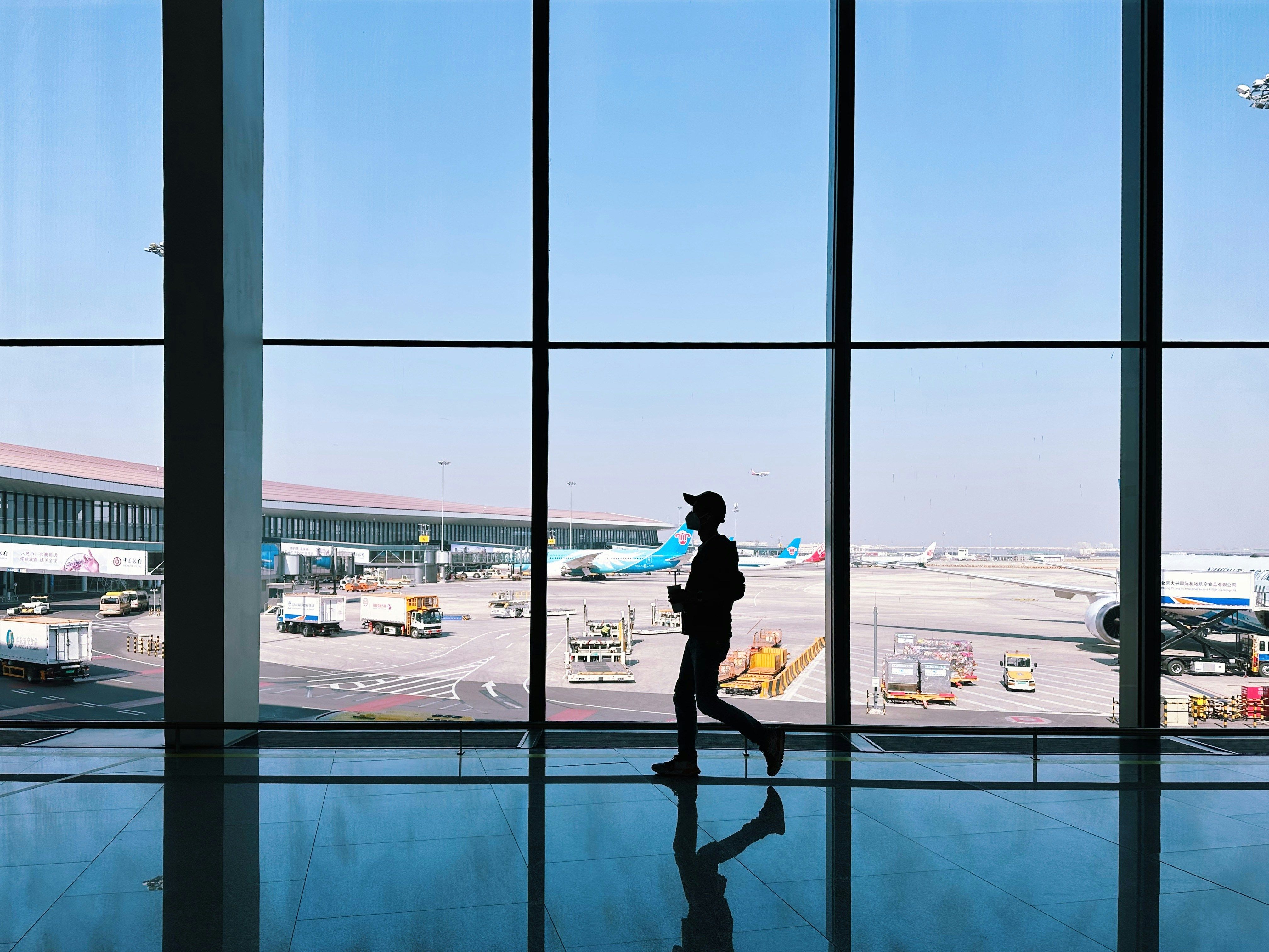 Silhouette of a traveler walking through an airport terminal with planes and ground vehicles visible outside the large glass windows.