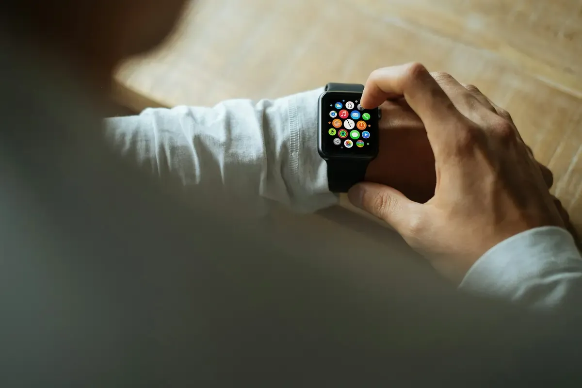 A person wearing a smartwatch interacts with the app grid on the watch screen while seated at a table.