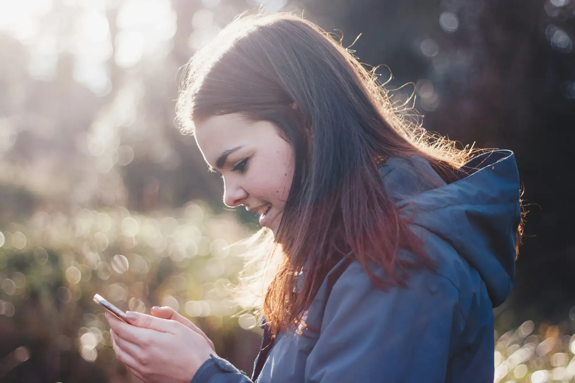 Traveler in a blue jacket using a smartphone outdoors, with sunlight filtering through trees in the background.