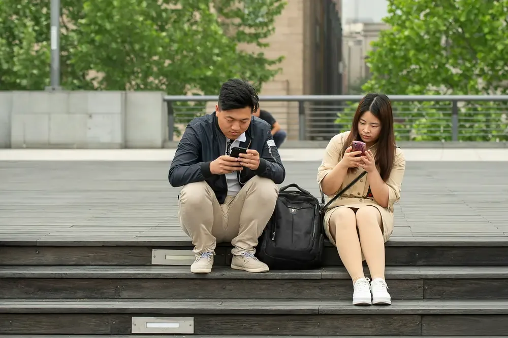 Two individuals focused on smartphones, sitting on outdoor steps.