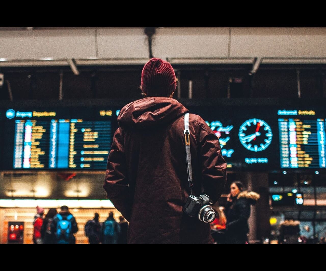 A traveler wearing a backpack and camera looks up at a departures board inside a busy airport terminal.