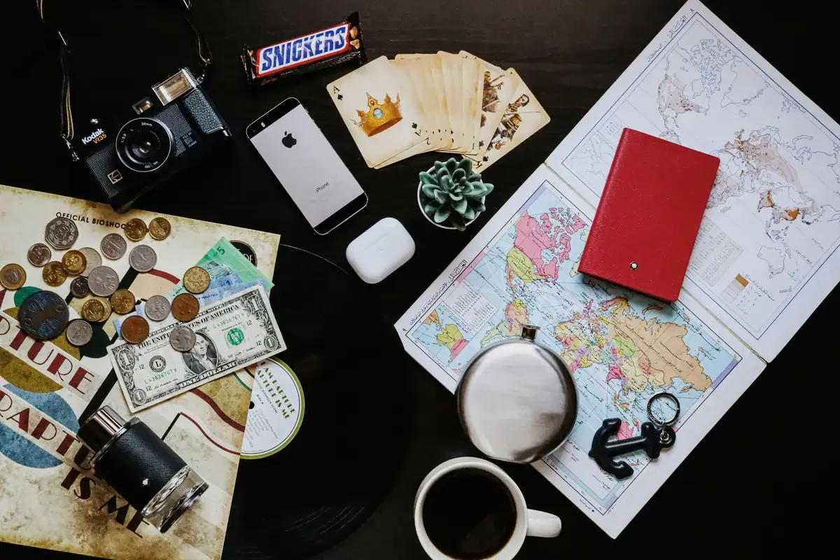 A flatlay of travel essentials including a vintage camera, world maps, foreign currency and coins, a passport, playing cards, an iPhone, earbuds, a Snickers bar, and a cup of coffee on a dark table.