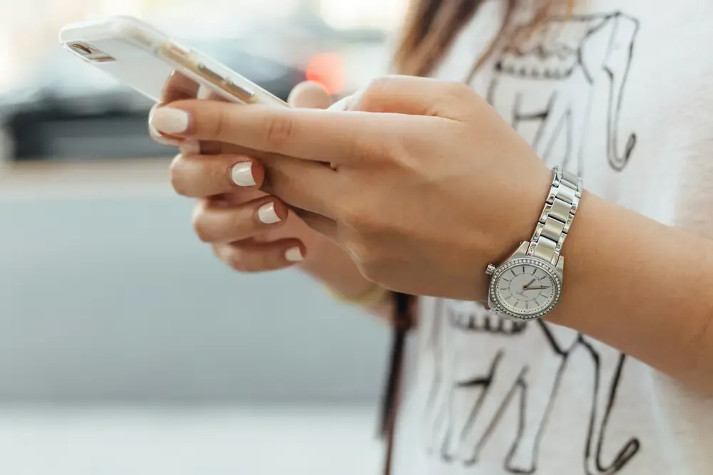 Close-up of a woman using a smartphone, wearing a silver wristwatch and a white shirt with an elephant print.