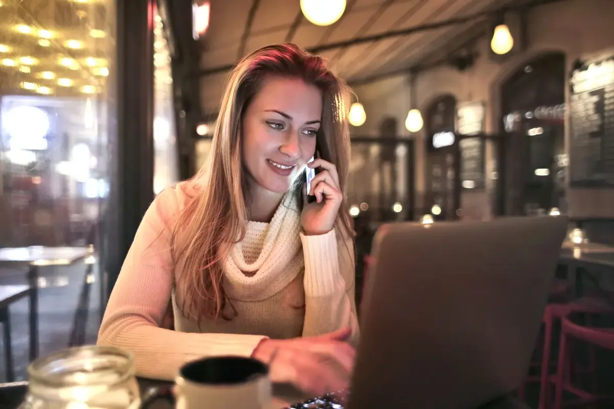 Woman on phone and laptop in a cozy cafe — representing VoLTE compatibility with eSIMs and mobile internet connectivity