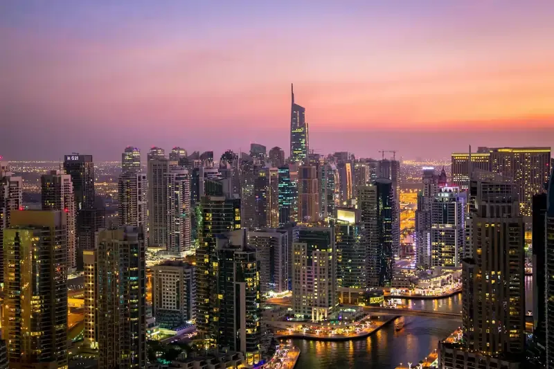 Skyline of Dubai Marina at sunset, featuring high-rise buildings, illuminated city lights, and reflections along the waterway.