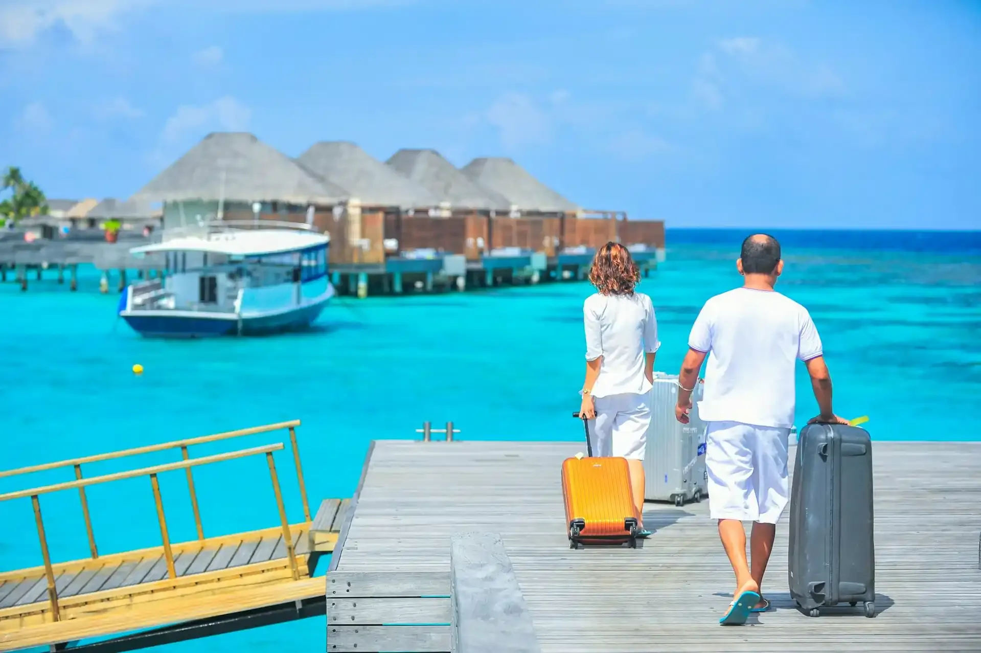 Couple walking on a pier with luggage toward overwater villas in a tropical resort.