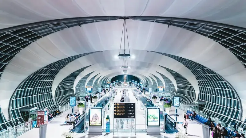 Wide-angle view of a modern, brightly lit airport terminal with arched architecture, central walkways, and travelers moving along travelators.