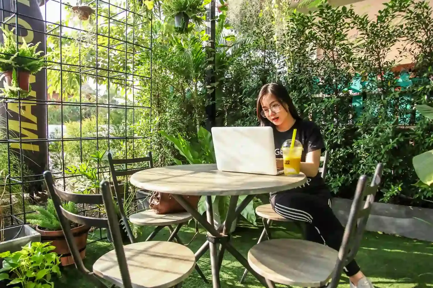 Young woman working remotely on a laptop at an outdoor café surrounded by greenery—ideal for digital nomads seeking nature and Wi-Fi.