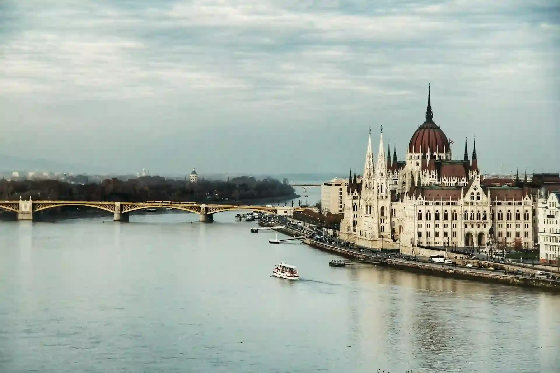Panoramic view of Budapest’s Parliament Building and the Danube River, with a sightseeing boat and bridge—an iconic and budget-friendly European travel destination.