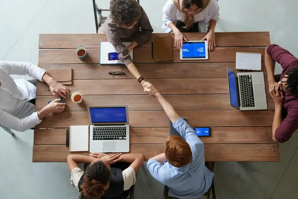 Overhead view of a business meeting with six people around a wooden table, using laptops and tablets, with two individuals shaking hands across the table.