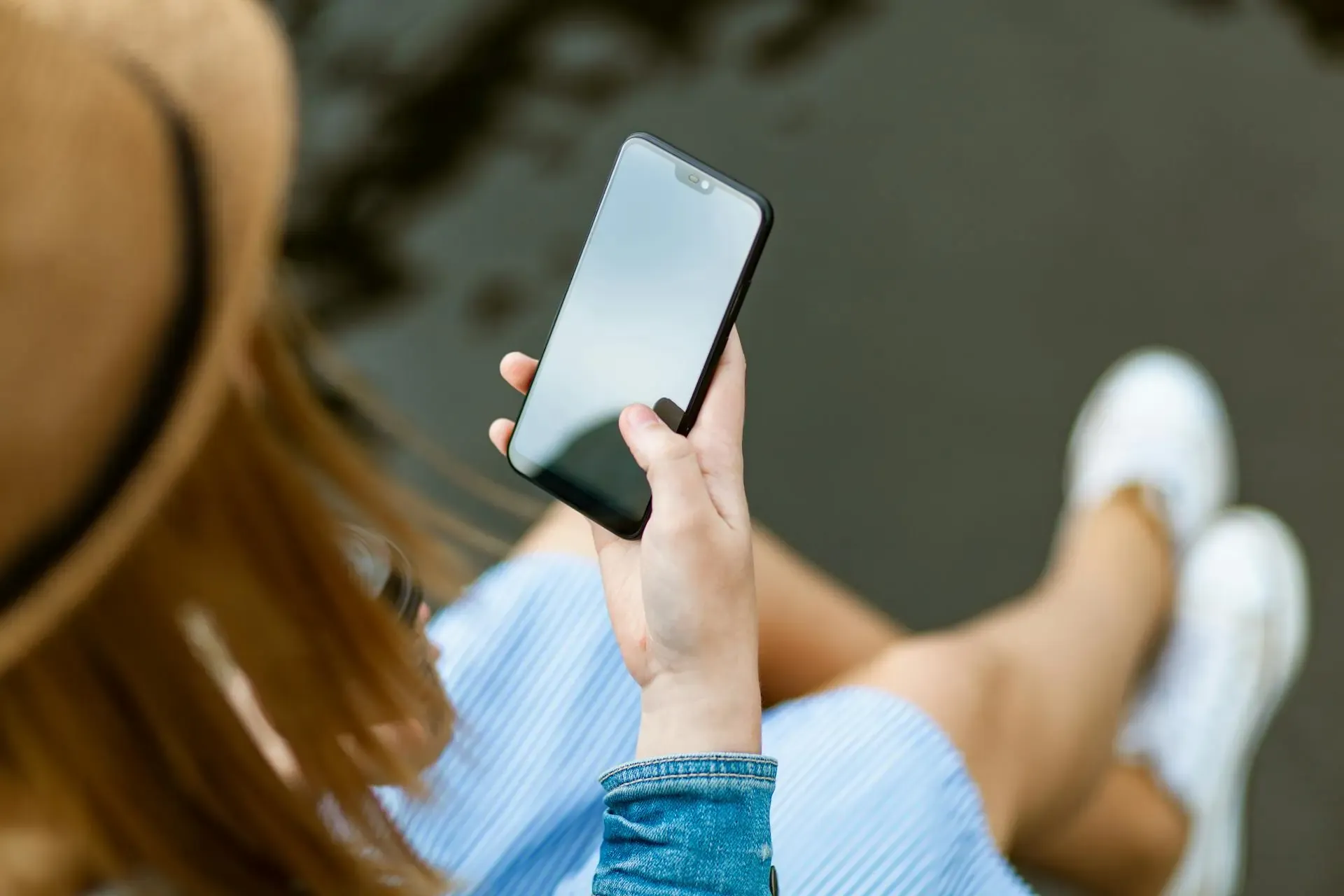 Over-the-shoulder view of a woman holding a smartphone with a blank screen, sitting outdoors in casual attire near water
