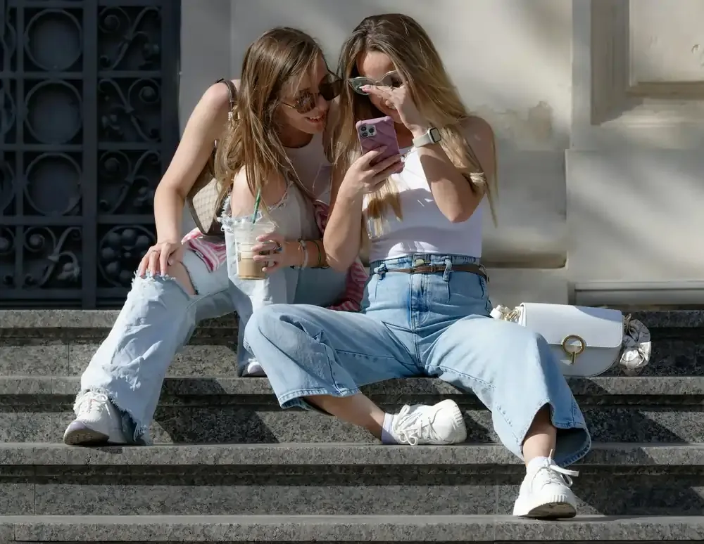 Two young women sitting on city steps looking at a smartphone, comparing international roaming and travel eSIM options.