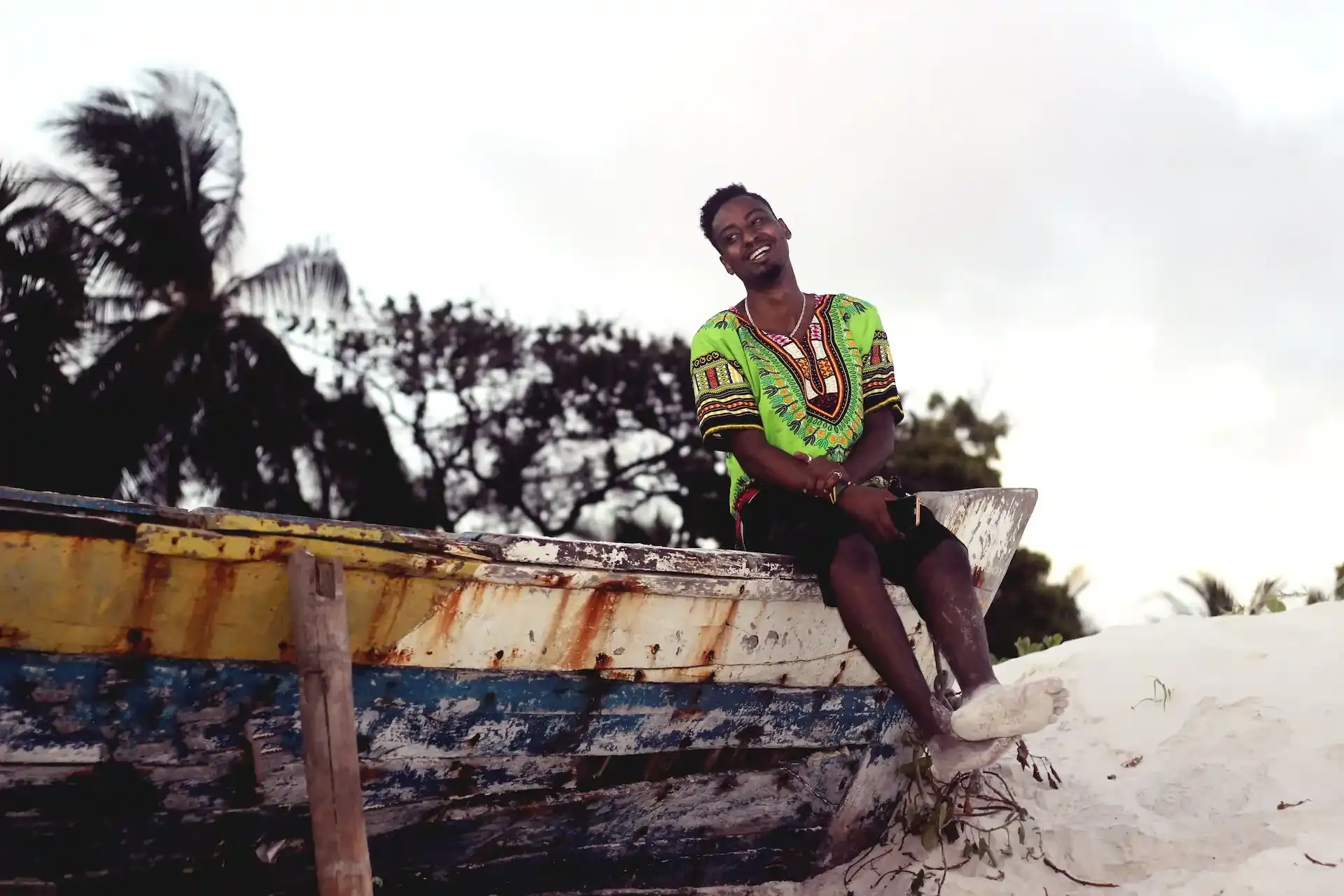 Smiling man in traditional Kenyan attire sitting on a rustic boat by the beach, capturing the relaxed vibe of travel in Kenya.