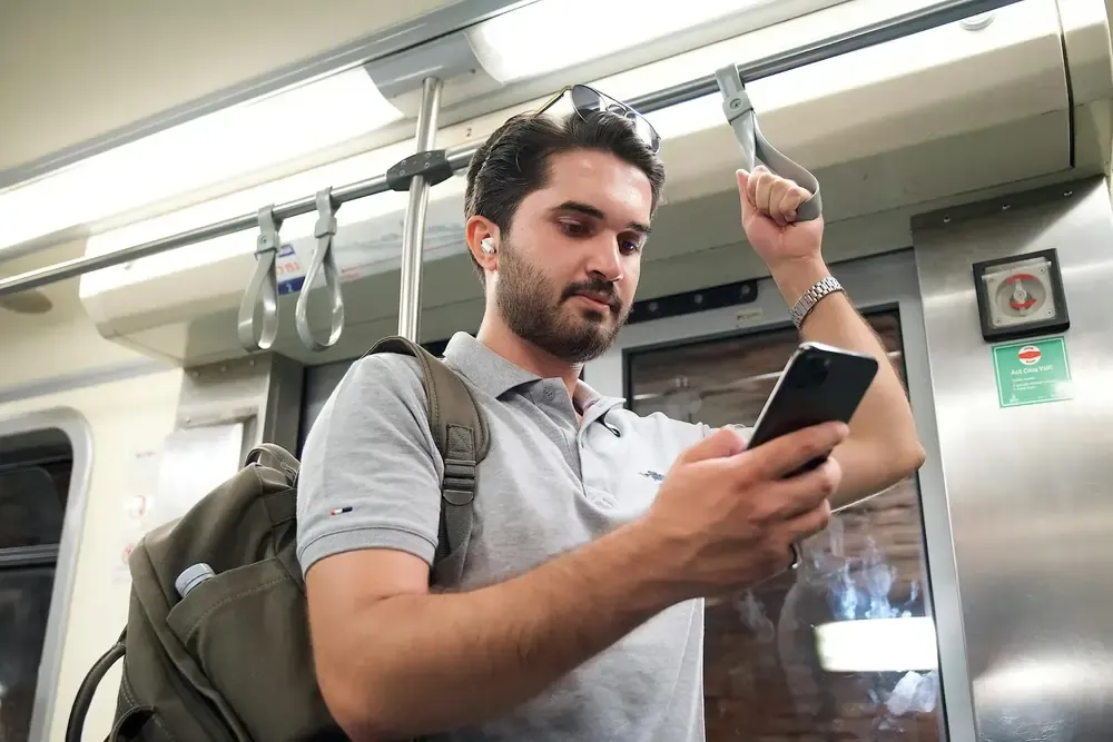 Man standing in a subway train, holding onto a handrail with one hand and using his phone with the other