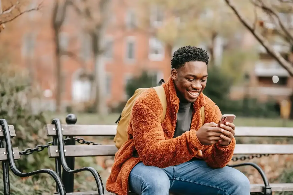 Man sitting on a park bench smiling while using smartphone, staying connected with eSIM during travel in autumn city setting.