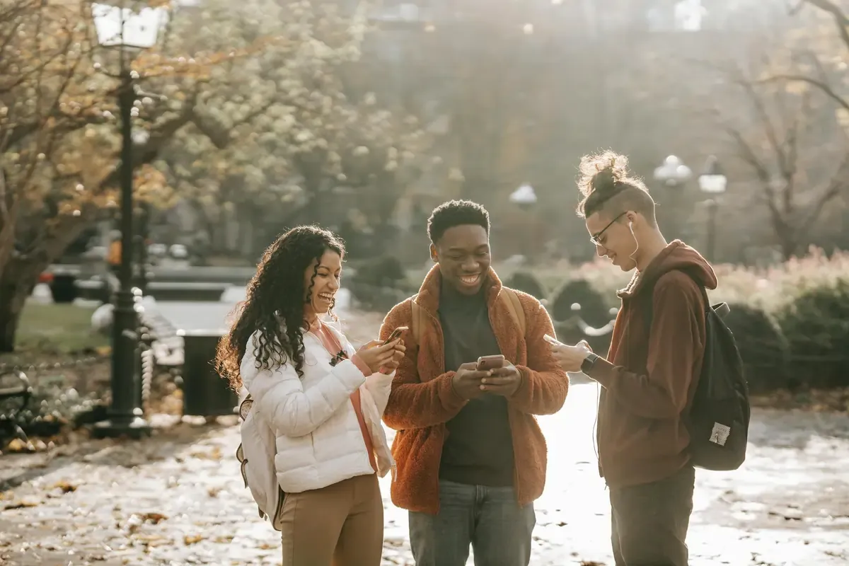 Three smiling friends using their smartphones outdoors in a sunlit park.