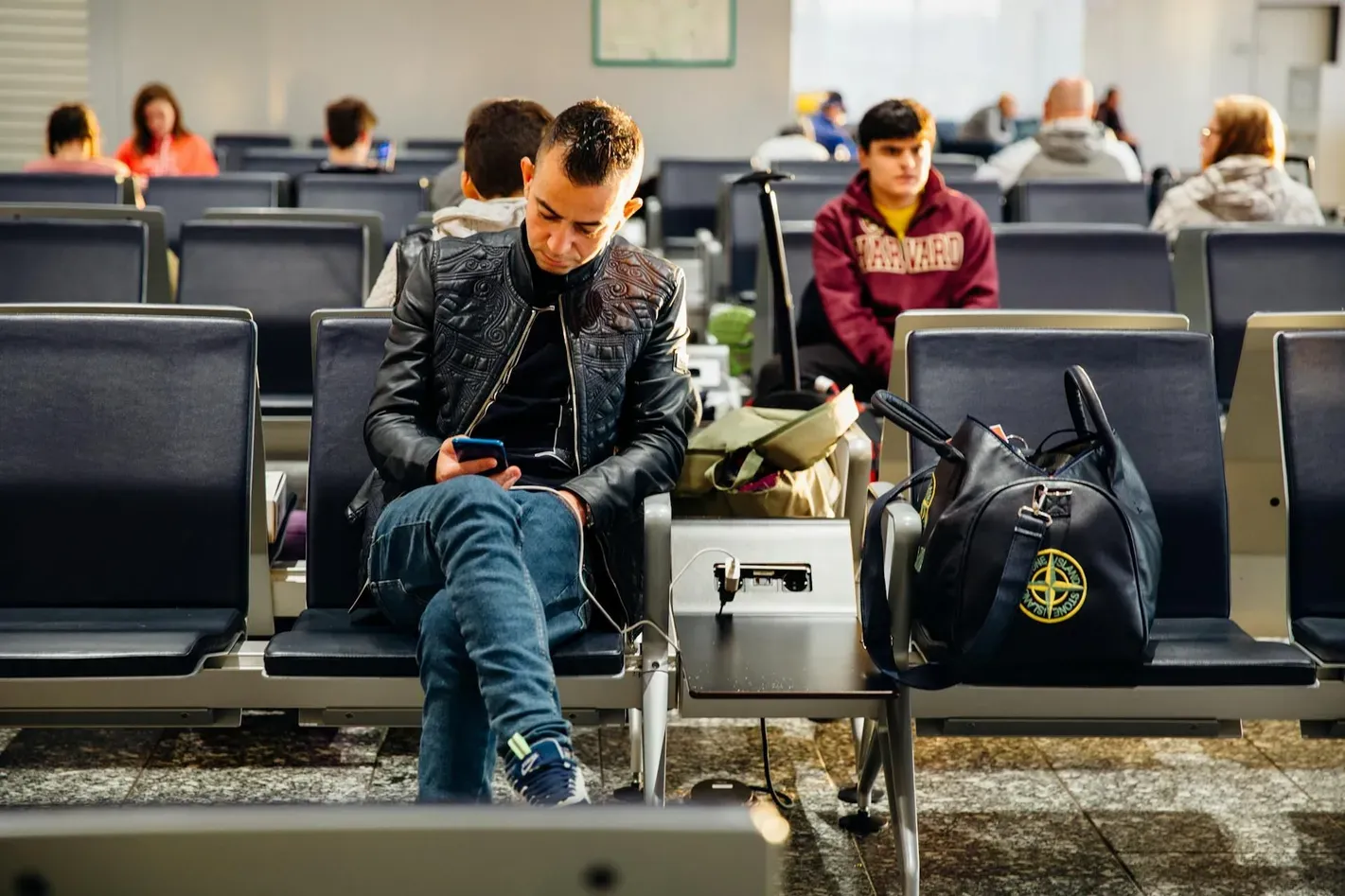 Traveler sitting in an airport terminal using a phone, with a carry-on bag beside him—illustrating essentials to pack in your carry-on for convenient and connected travel.