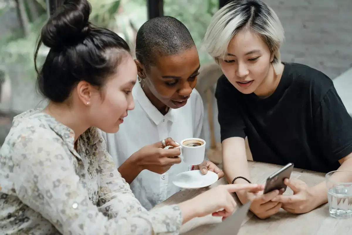 Three women sitting at a table in a casual setting, looking at a smartphone together.