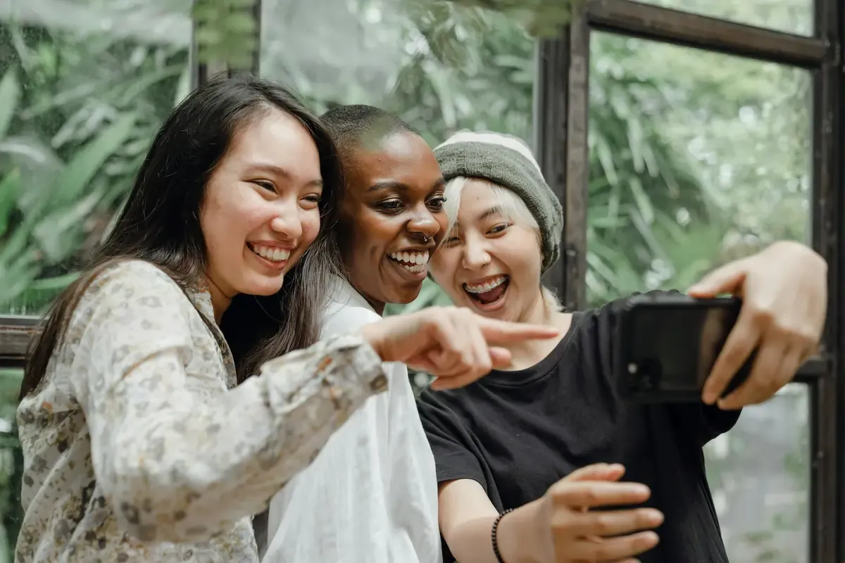 Three friends smiling and laughing while taking a selfie together, capturing a joyful moment indoors surrounded by greenery.